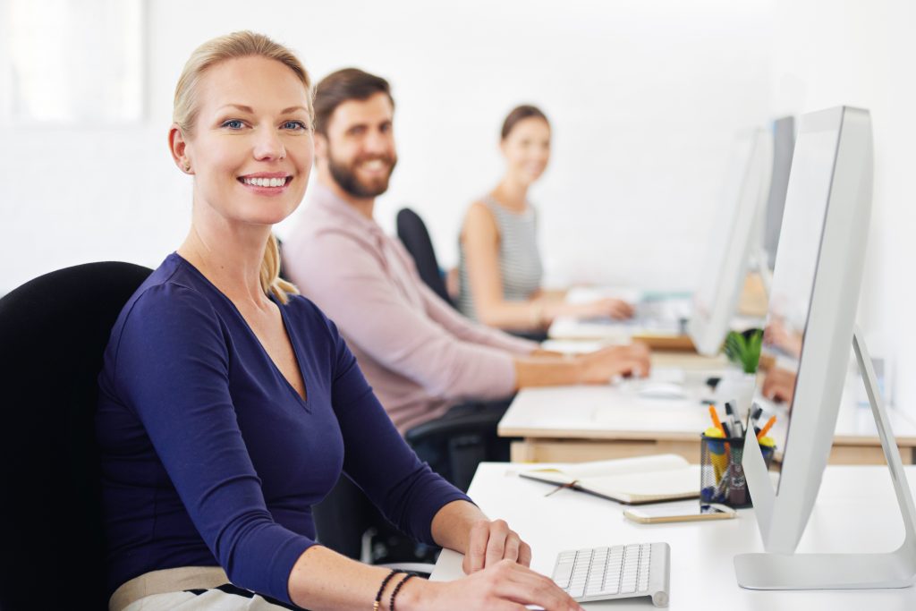 3 Personen arbeiten an Computern an einem hell ausgeleuchteten Schreibtisch.